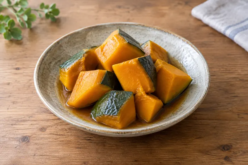 A small ceramic bowl of glossy simmered kabocha chunks, bright orange flesh with patches of dark green skin, sitting in a little of their amber cooking liquid