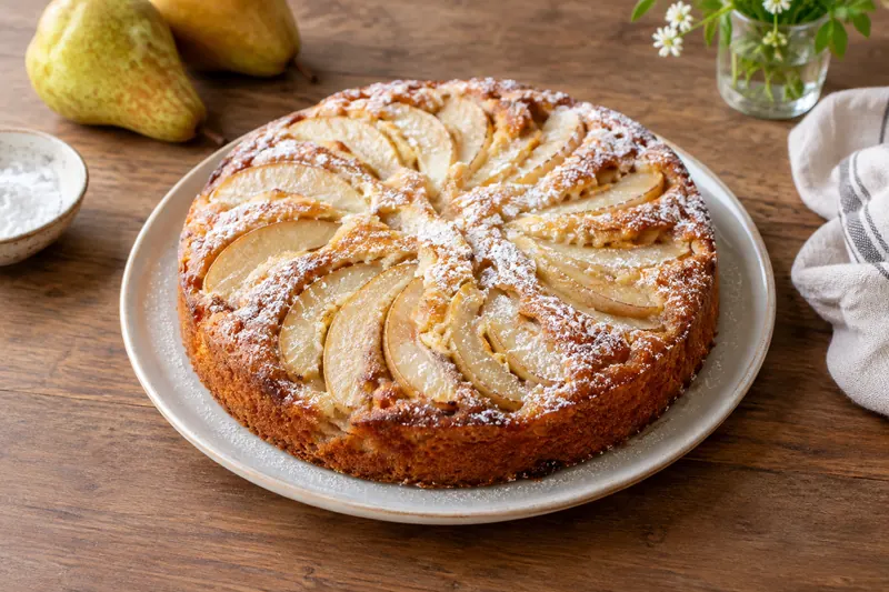 A round golden pear cake on a plate, its top covered in fanned baked pear slices and dusted with icing sugar, one slice cut out.